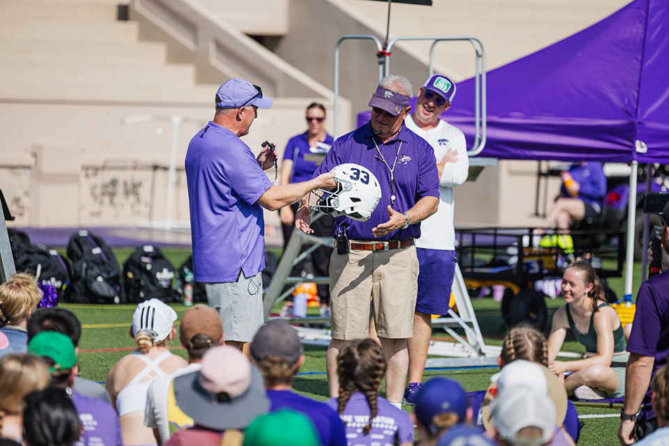 A football coach in a purple polo hands a signed helmet to a band director out on a practice field.