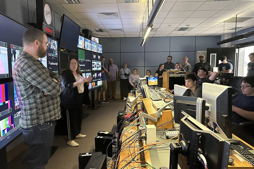 A duo of news producers stand in front of a television production suite and present to a group of college students sitting at the suite's stations.