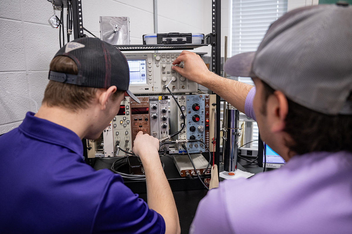 Two young men stand in front of a panel with a lot of different knobs and buttons on it.
