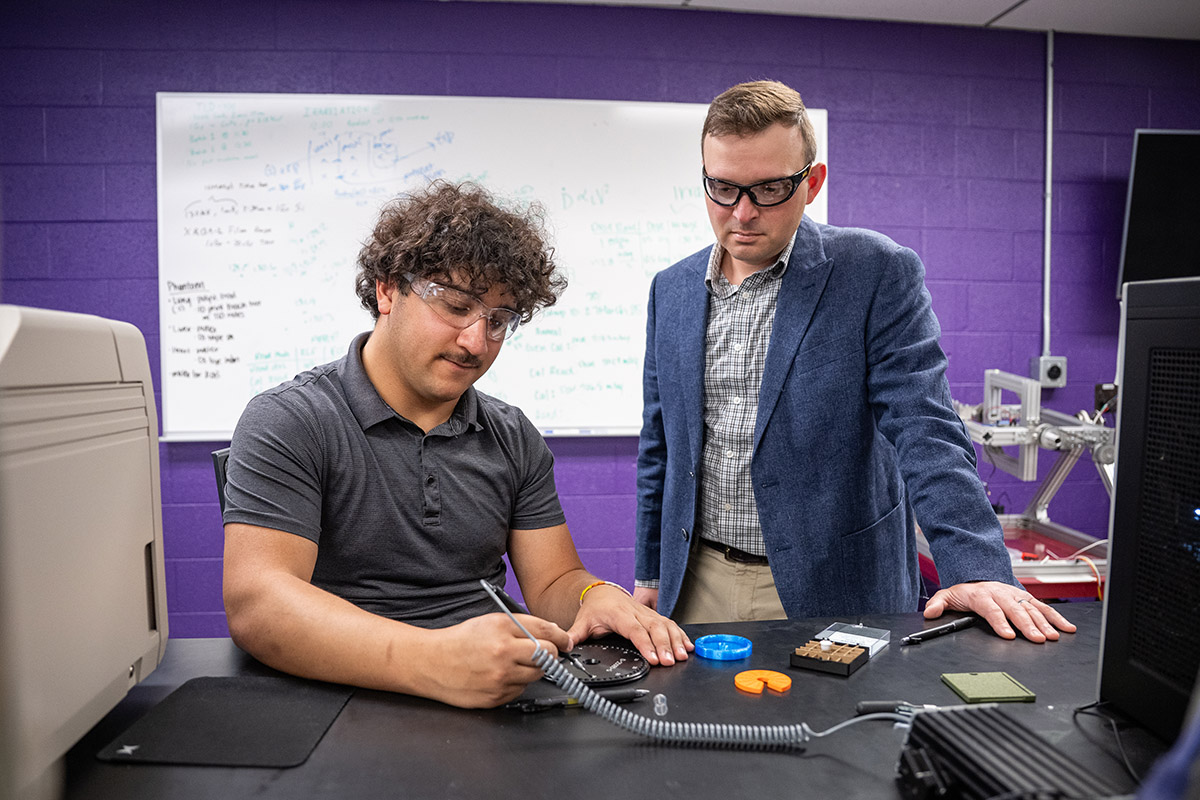 A young man in safety googles sits at a desk working with equipment while a professor in a suit coat and safety goggles watches intently.