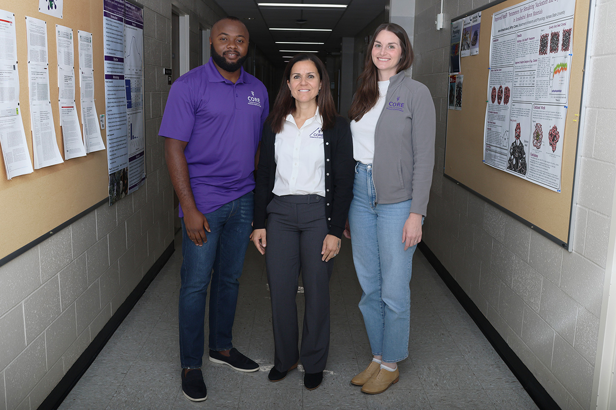 A Black man with a beard, a woman with brown hair, and a woman with dark hair stand in a hallway.
