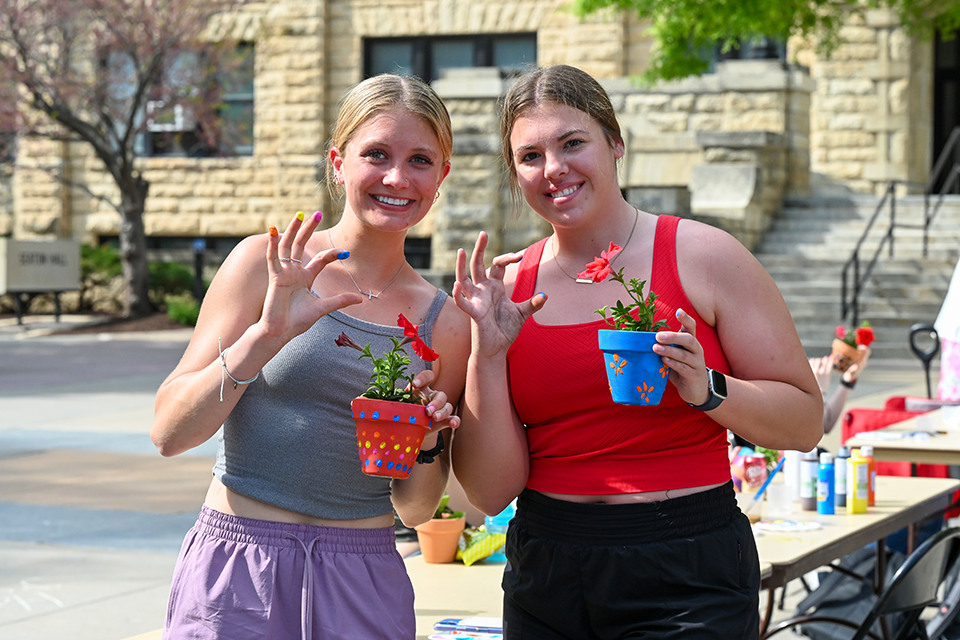 Two female college students stand outside of a limestone building while each holding decorated flowerpots and making the Kansas State University Wildcat hand symbol. 