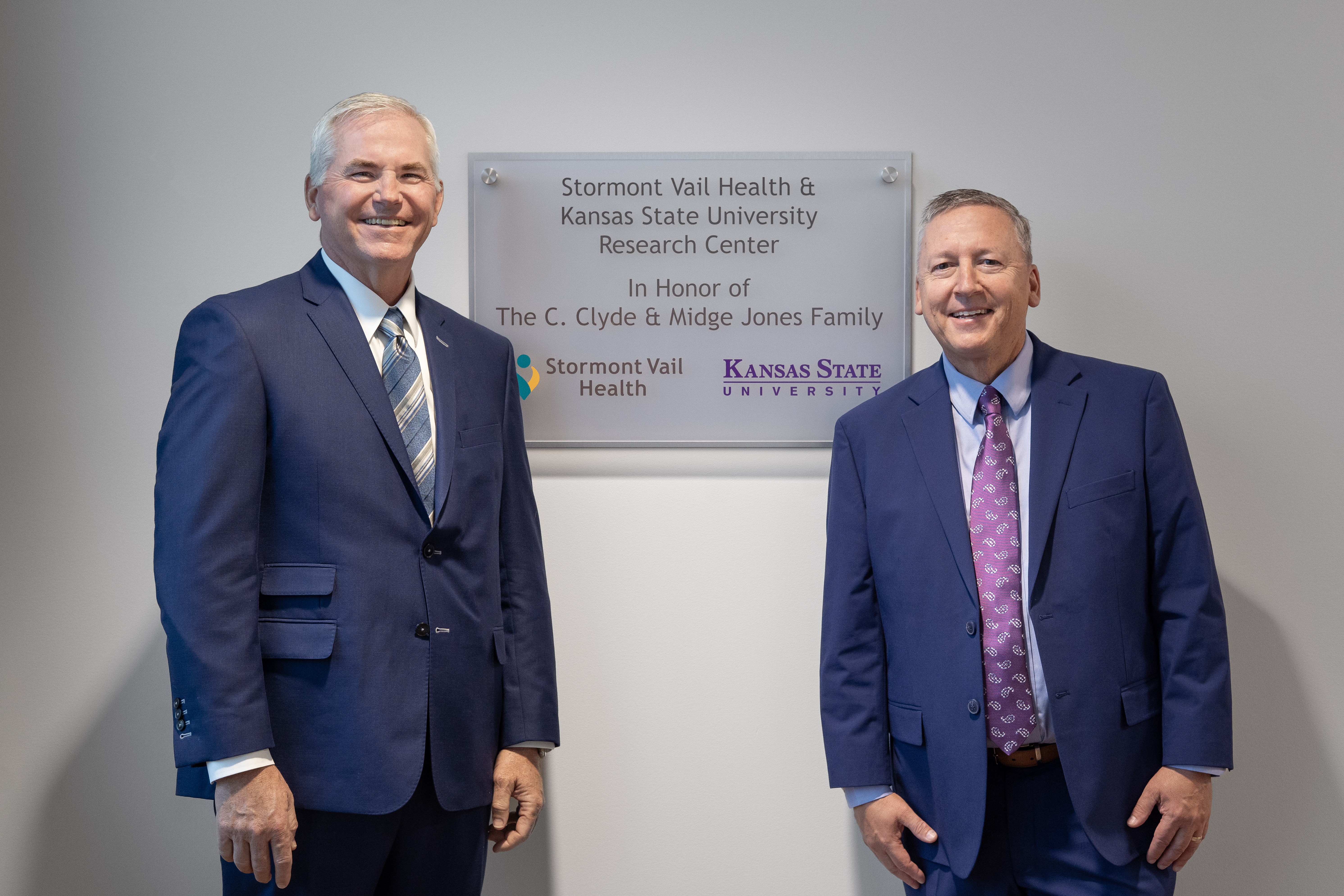Two men in navy suits stand in front of a glass panel on a wall that says, "Stormont Vail Health & Kansas State University Research Center in honor of the C. Clyde and Midge Jones Family"