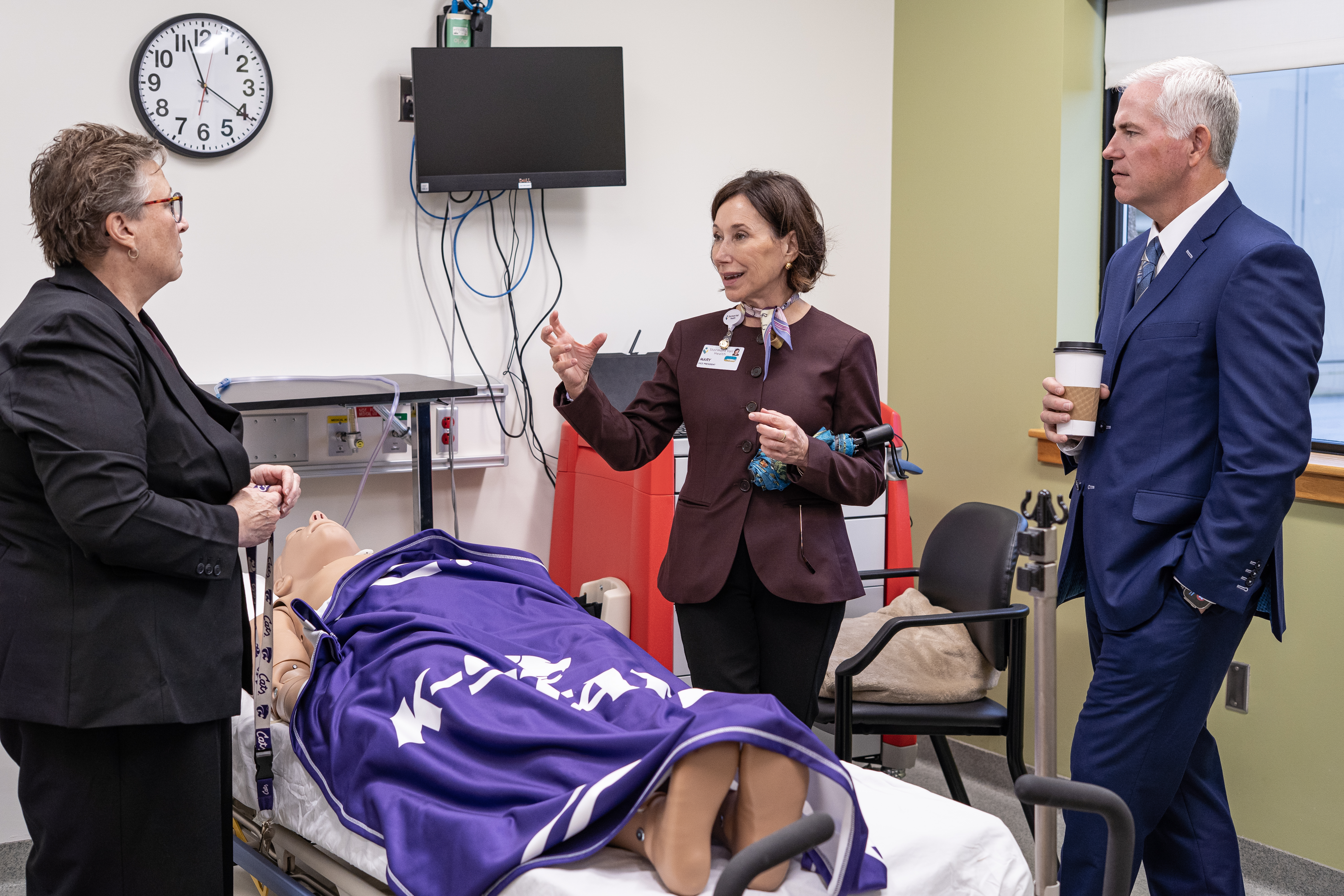 Three people in professional clothes stand near a mock hospital setup with a health mannequin on a bed covered in a purple K-State blanket. A woman in a mauve professional shirt wearing an ascot gestures to a woman in a gray suit jacket and glasses across the bed while a man in a navy suit listens.