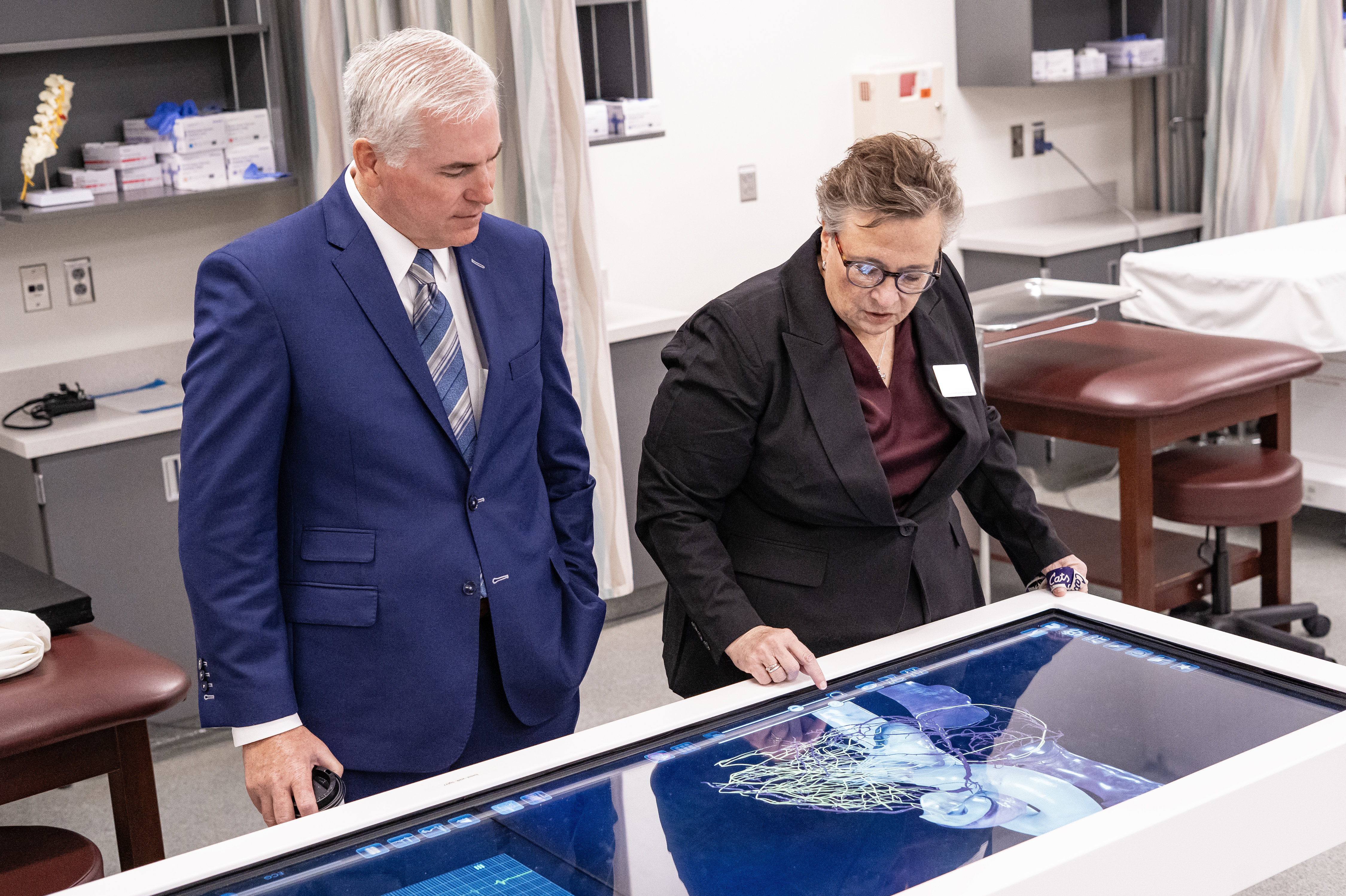 A man in a navy suit and a woman in a gray suit look at and interact with a digital screen.