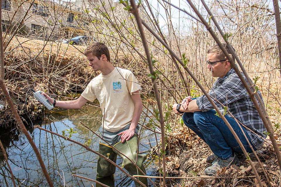 A college professor crouches next to a stream as a student pulls in a water sample from it on a college campus.