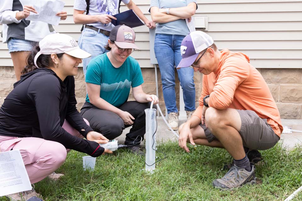 A college professor crouches and coaches college students on how to collect groundwater samples on a lawn outdoors.