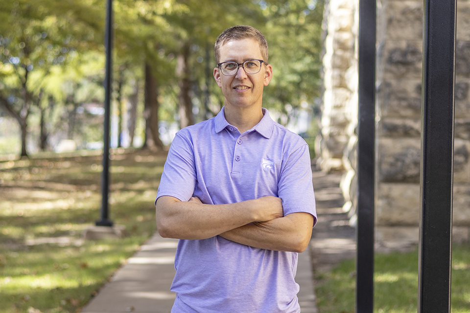 Matthew Kirk crosses his arms and poses for an outdoor portrait on a college campus.