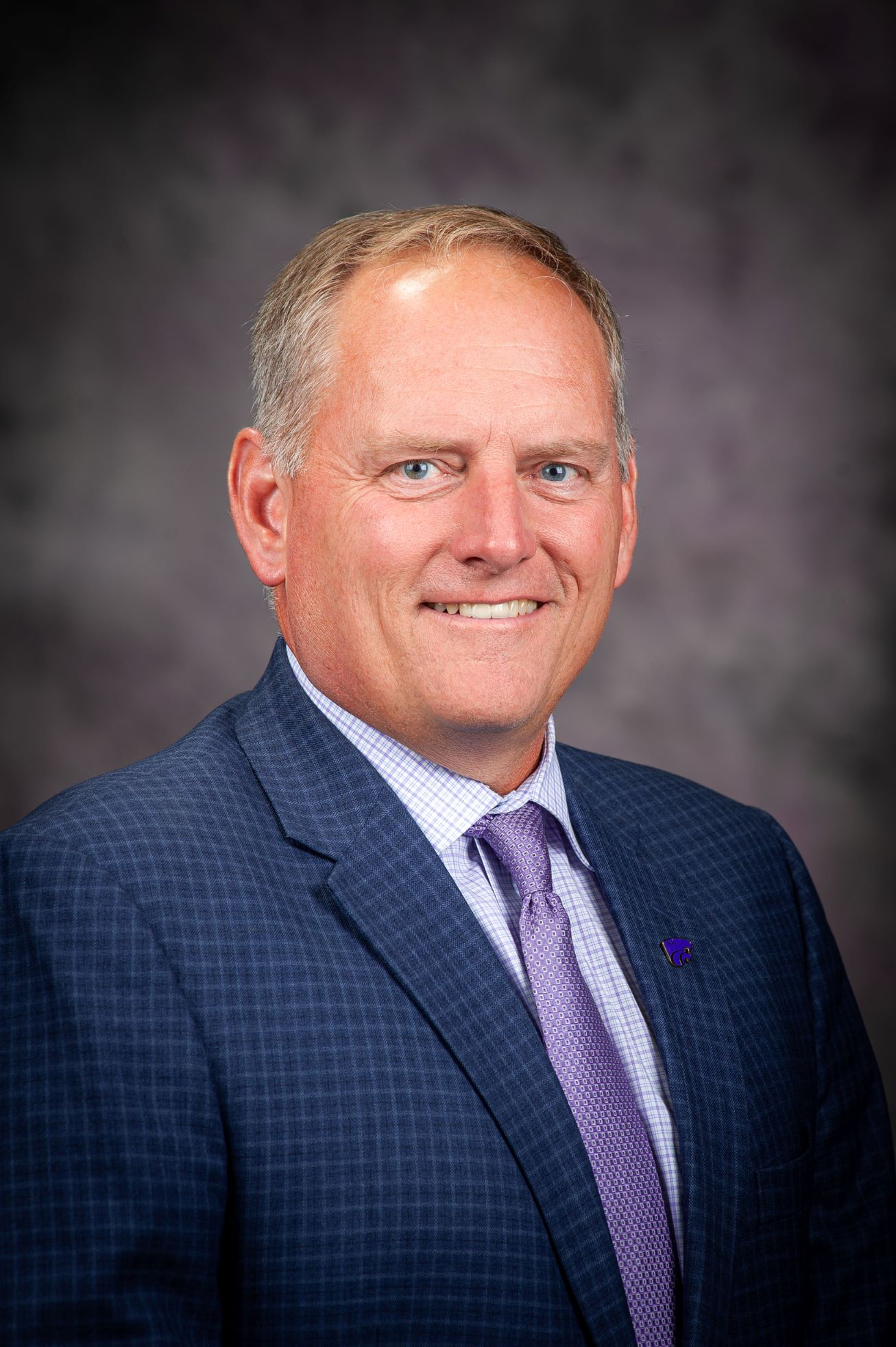 A man in a blue suit smiles for a studio portrait. 