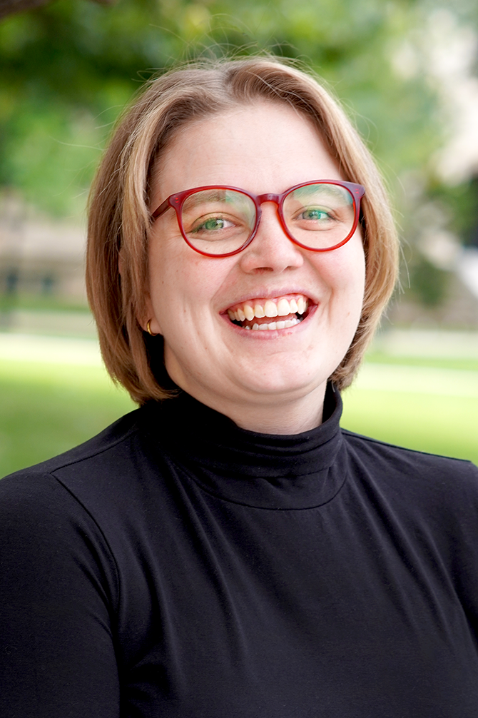 A doctoral student with short blonde hair and red glasses wears a black turtleneck and laughs in an outdoor portrait with greenery in the background.