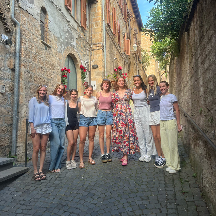 A group of students and a faculty member stand in an Italian alleyway surrounded by buildings, flowers and greenery.