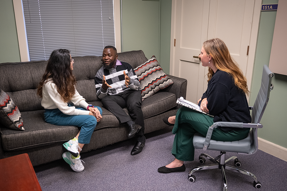 A couple sits on a couch in an office talking while a therapist sits in a chair across from them with a notebook and listens. 