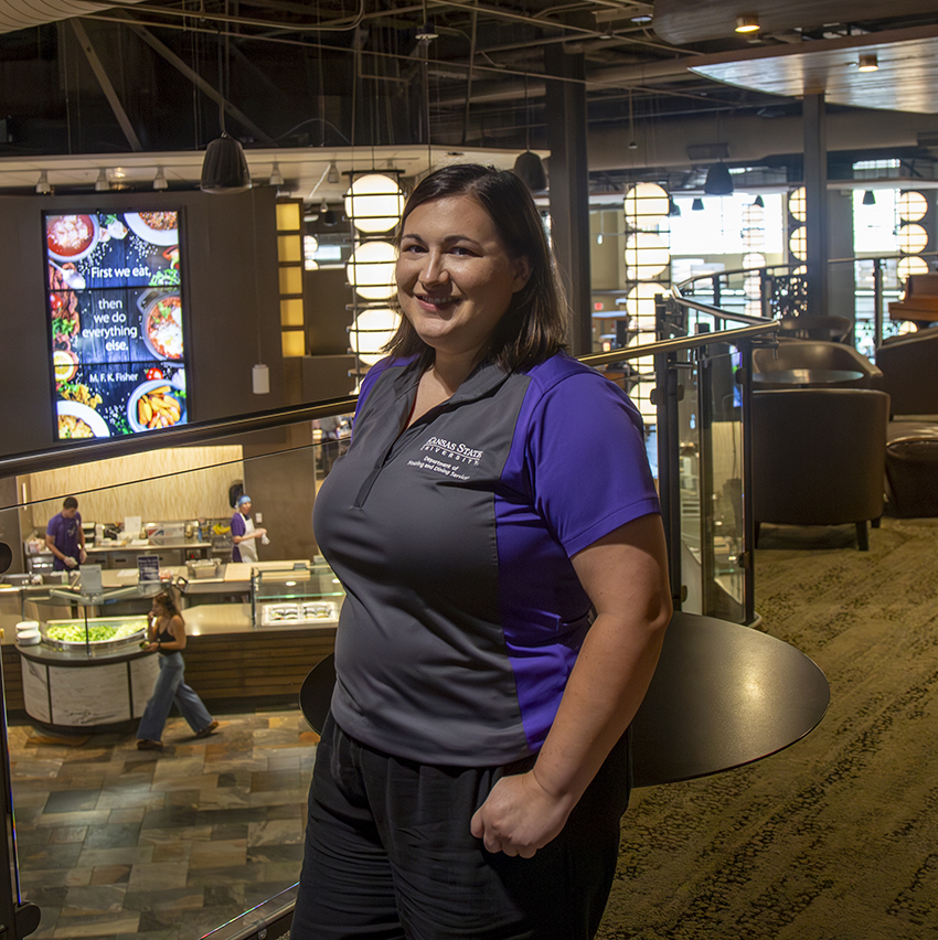 A woman in a purple polo poses for a portrait on a walkway above a college dining center.