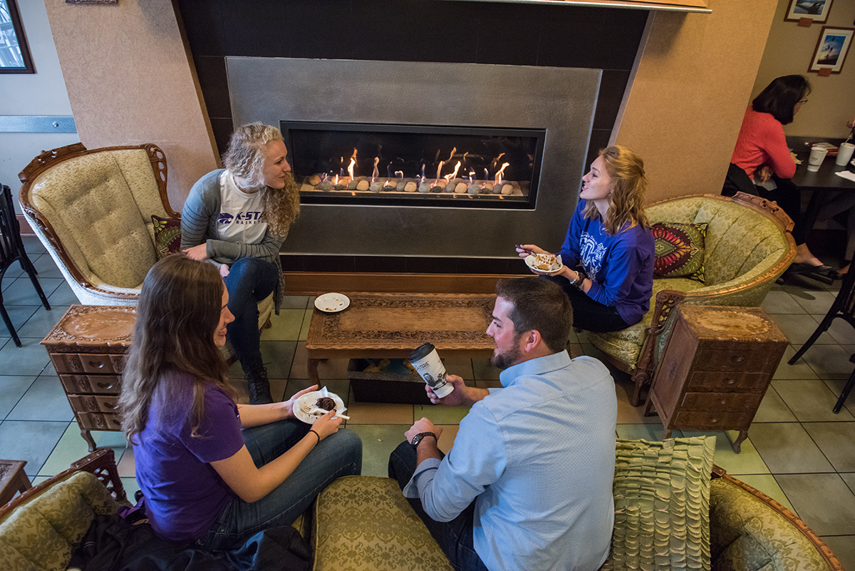 Four students sit in comfortable chairs near a fireplace. Two of the students hold plates with pastries on them, and one holds a to-go coffee cup.