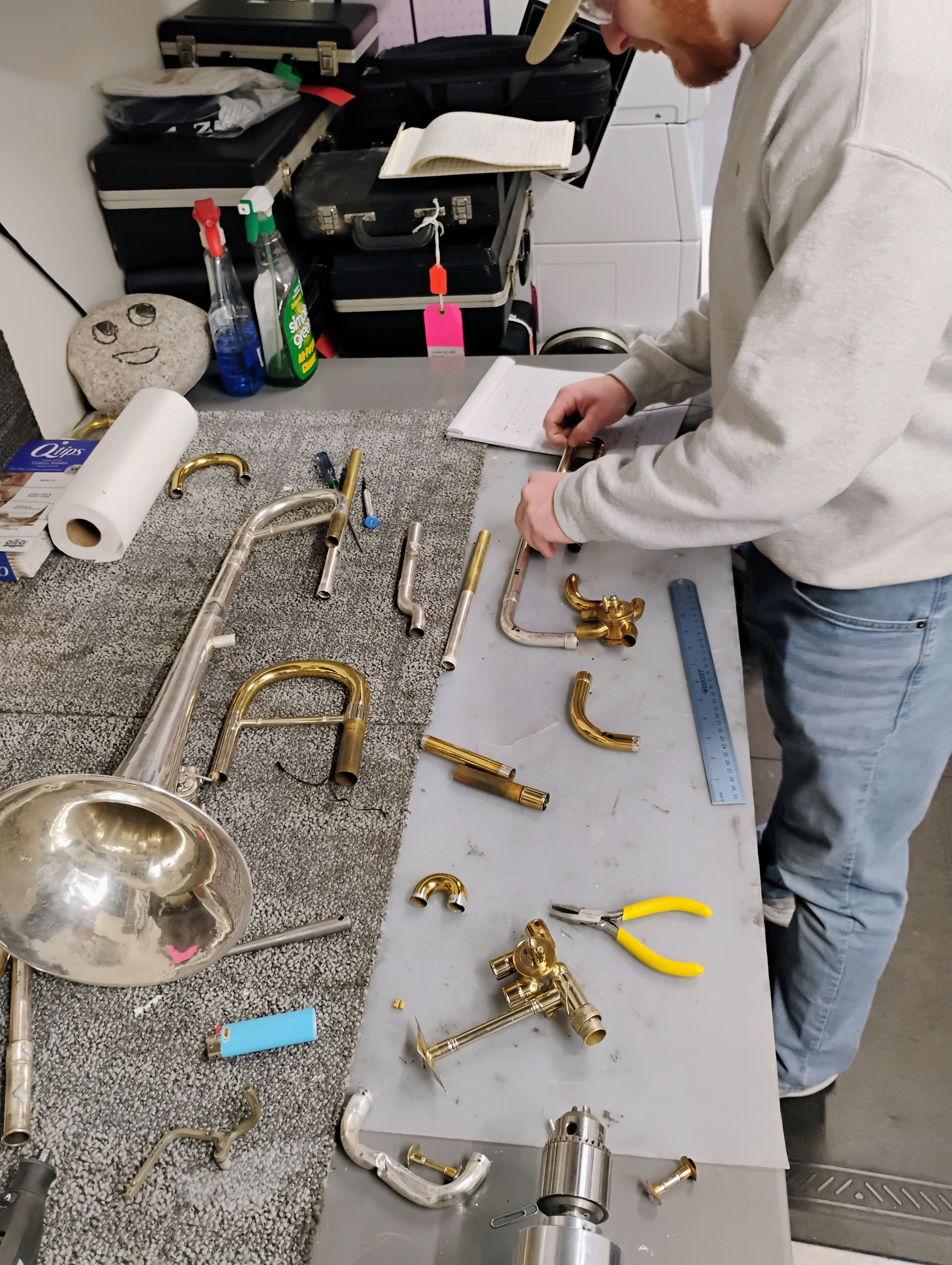 A student in a white sweater works on pieces of a trombone that are spread across a table. Also on the table are pliers and a metal ruler..