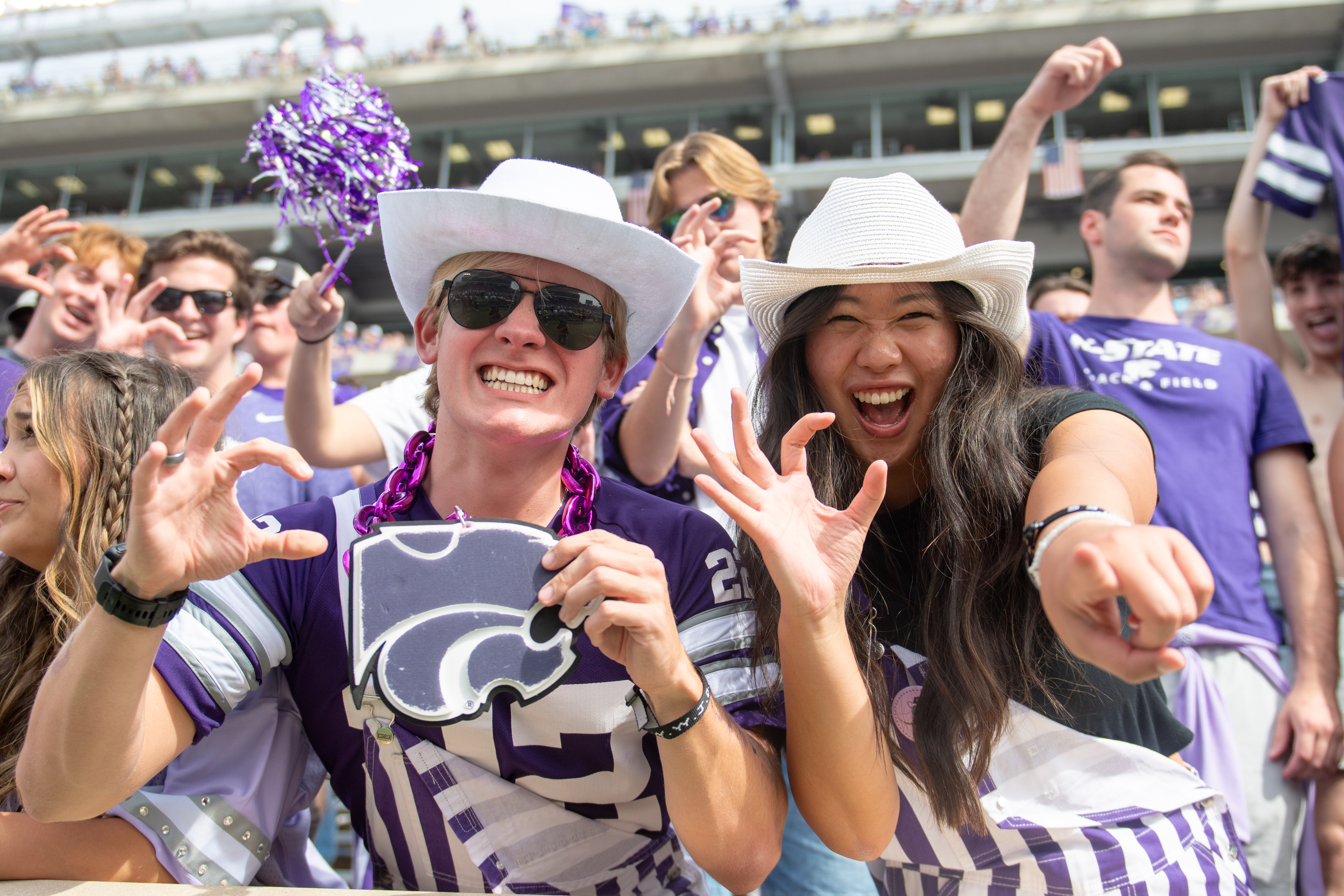 Two students in white cowboy hats and dressed in purple and white overalls make the Wildcat hand symbol and smile in the student section at a K-State football game. Other students are seen in the background making the hand symbol and cheering.