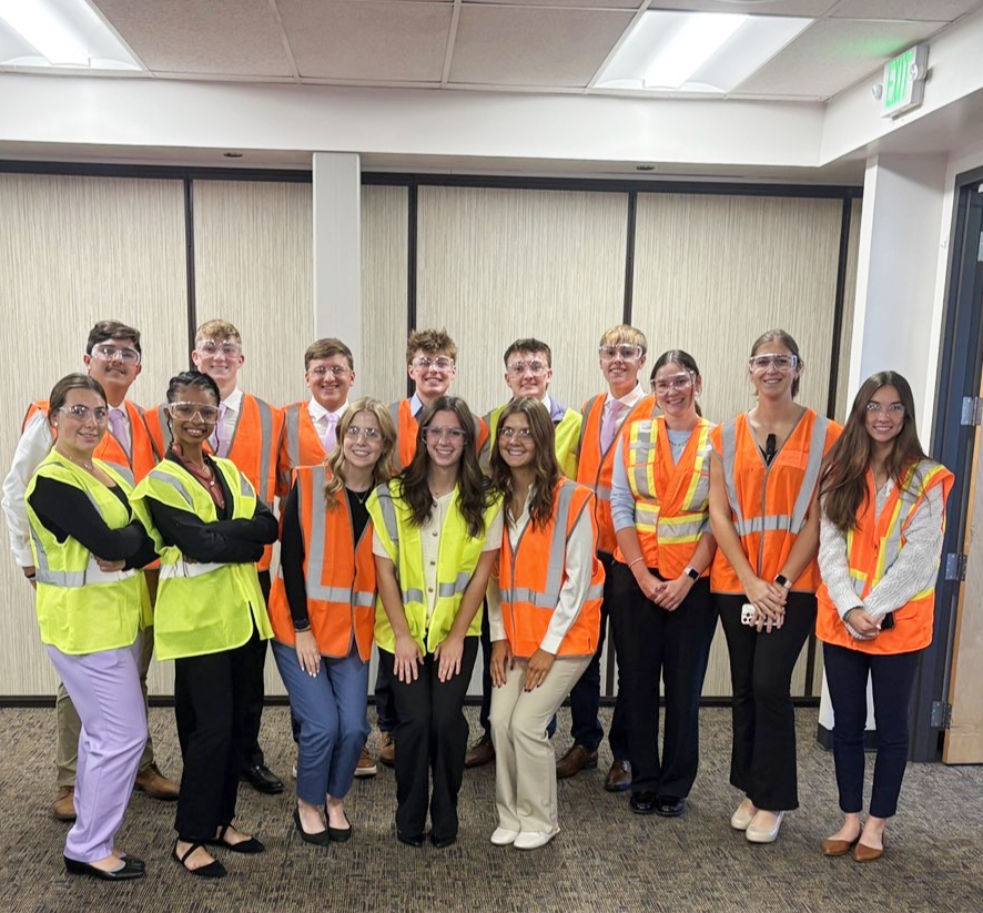 Fourteen students in orange and yellow safety vests and clear safety glasses stand in a line in an indoor room and smile for a group photo.
