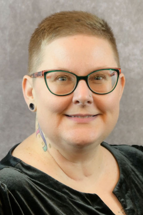 A staff member with buzzed blonde hair, glasses and black earrings smiles for a close-up portrait photo.