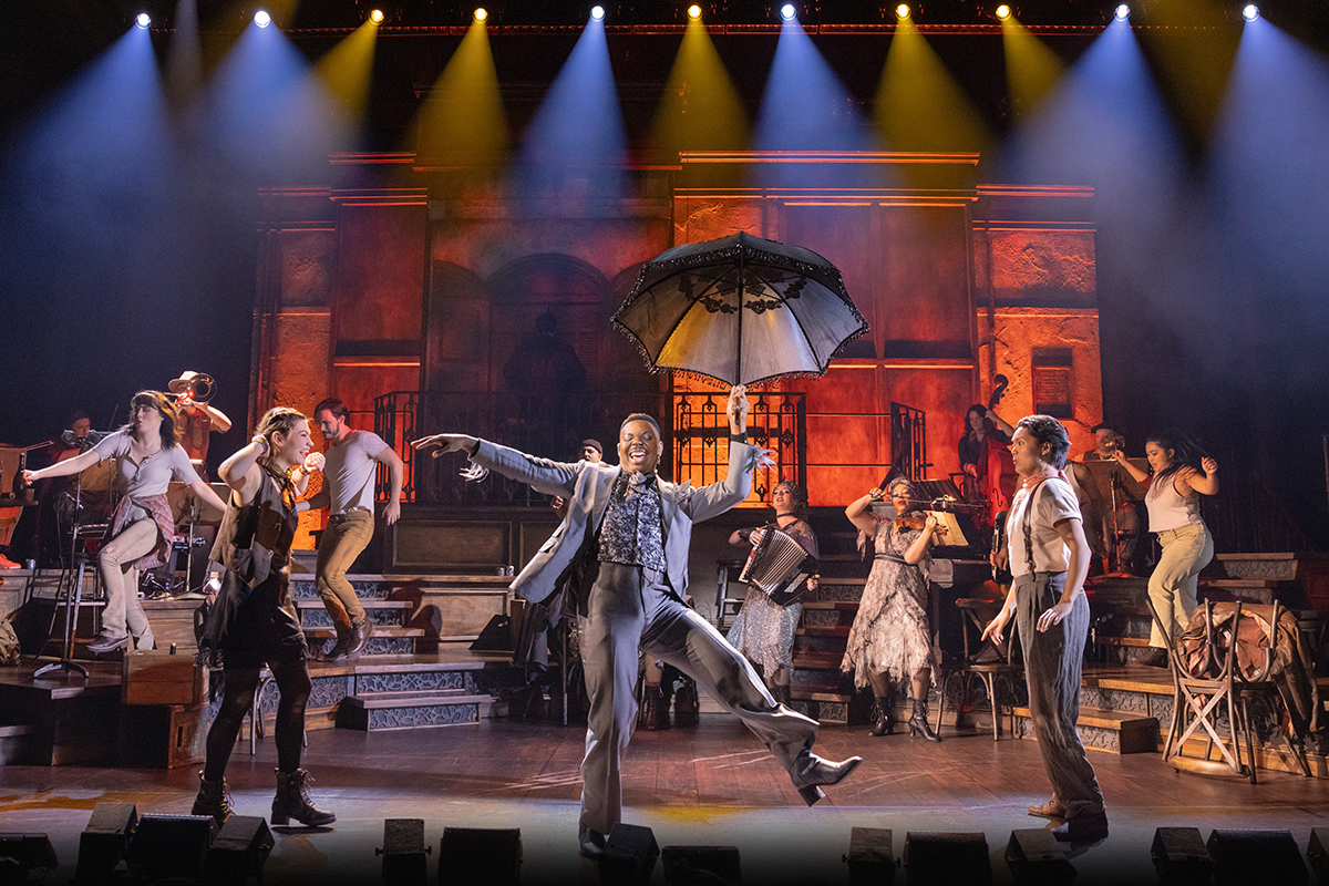 In a production of the musical Hadestown, a Black man in a gray suit holds a black umbrella onstage with a character on each side of him. In the background, various dancers and musicians are shown performing. Yellow and blue stage lights shine down from above.