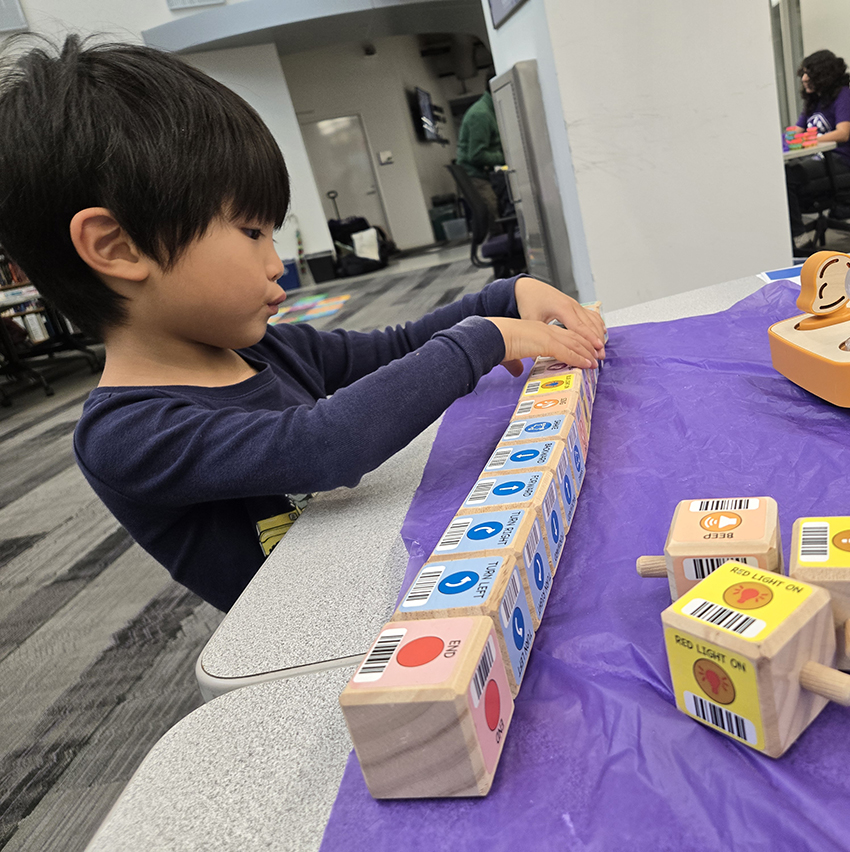 A young child concentrates as they line up wooden coding blocks on a table during a hands-on Hour of Code activity station.