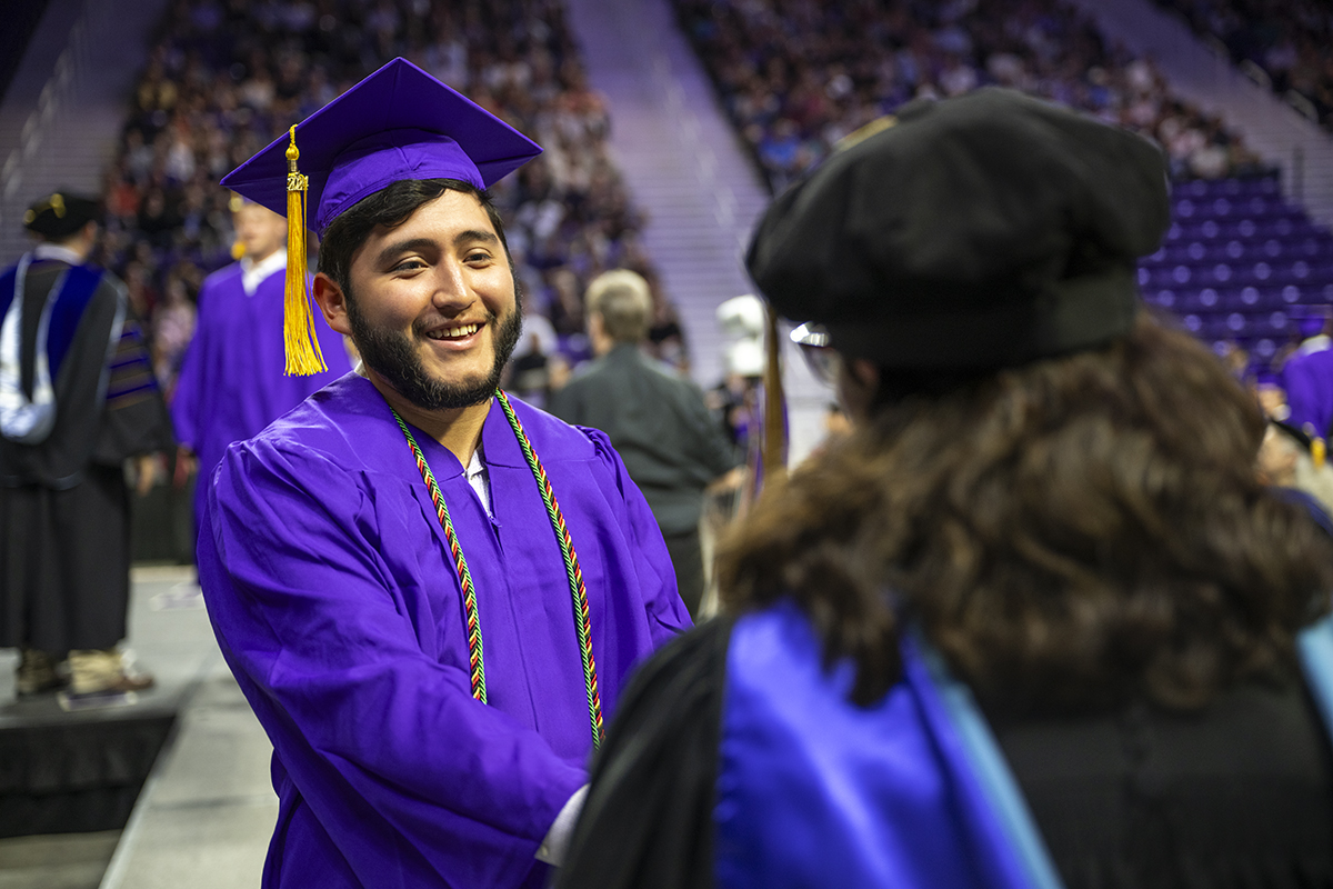 A college student in a purple cap and gown shakes the hand of a professor in graduation regalia.