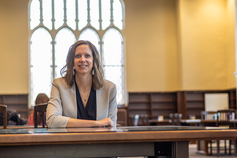 A woman sits at a table in a library and smiles for a portrait.