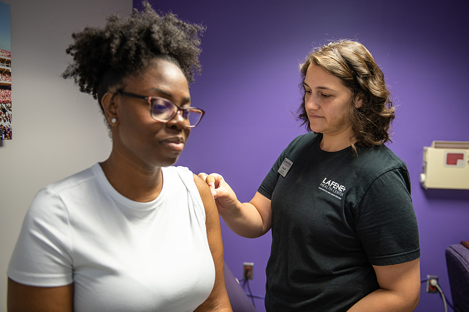 A close-up shows a pair of gloved hands injecting a flu vaccine into a person's arm.