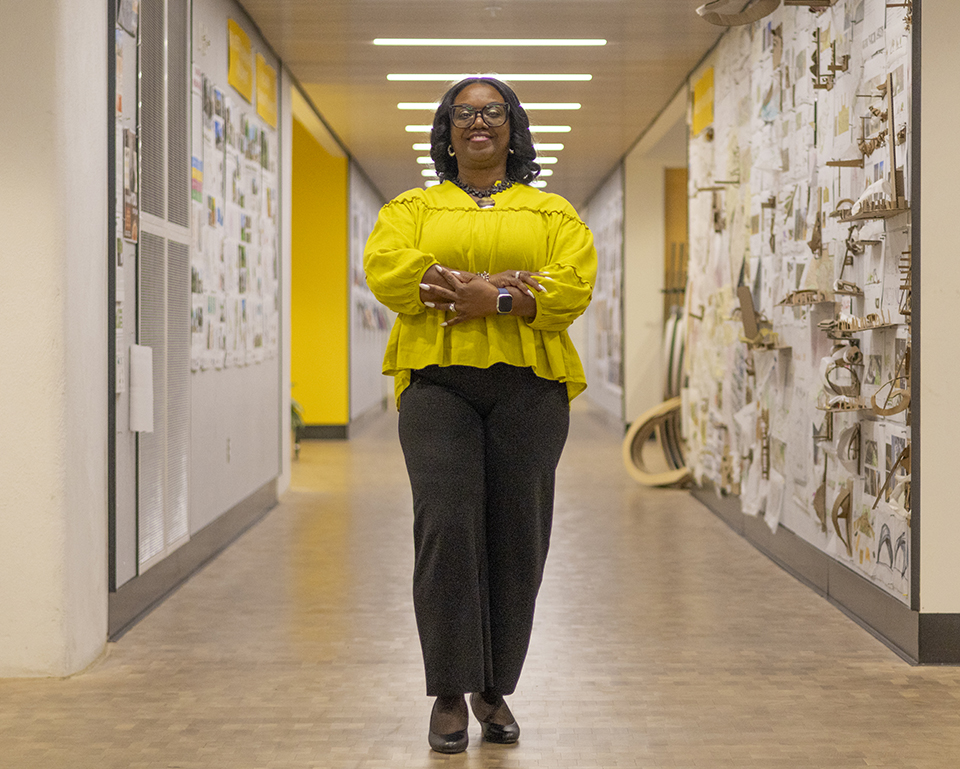 College advisor Mitzi Farmer crosses her arms in front of her and stands for a portrait in the center of a long college hallway.
