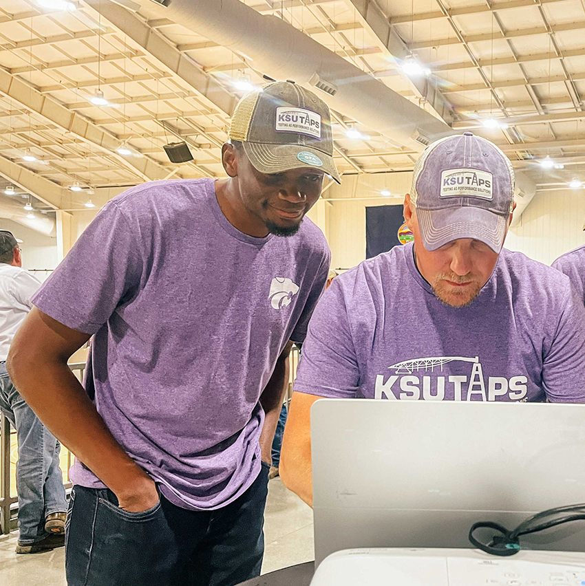 A graduate student in a purple T-shirt and hat stands and watches his professor type on a computer inside a large barn arena.
