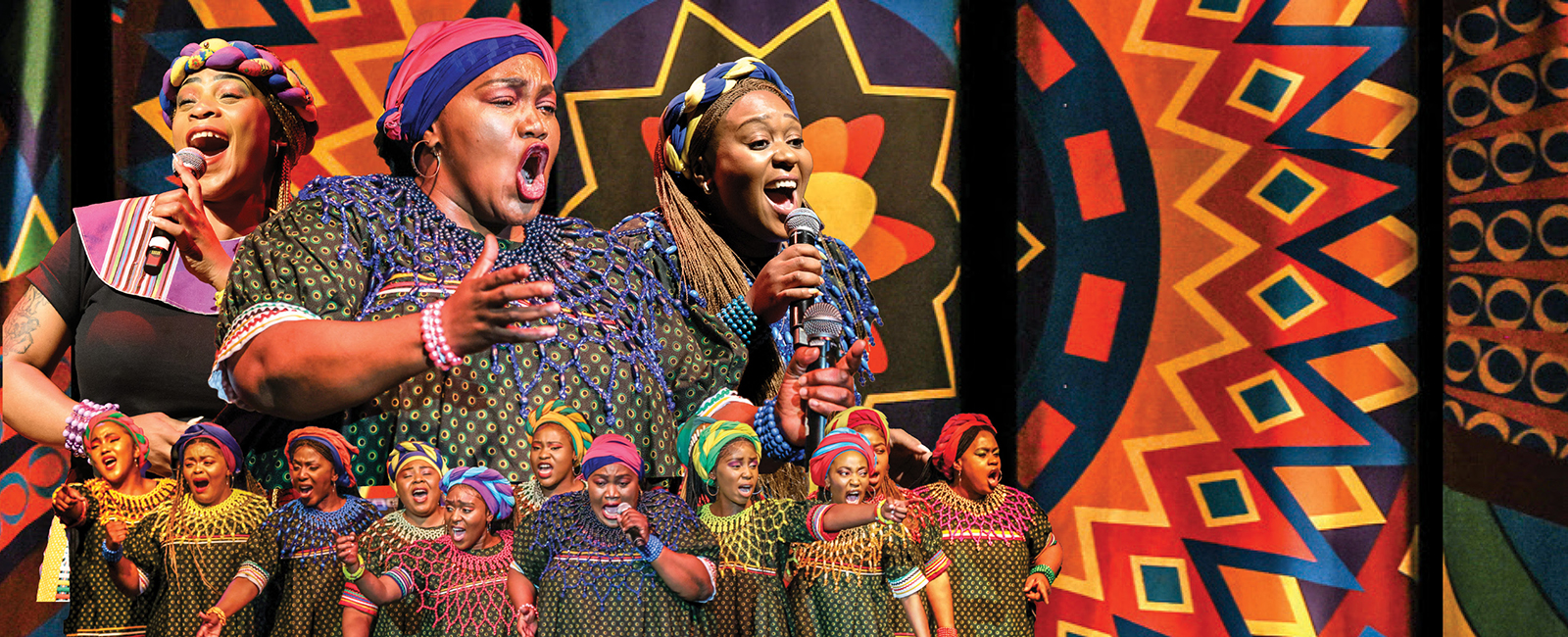 A group of women in African cultural attire sing on a stage.