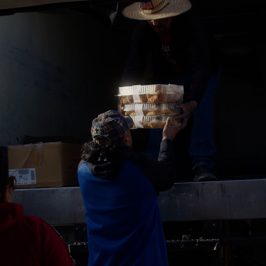 A person with long dark hair wearing a blue sweatshirt and a multicolored hat is handed multiple boxes of bread food from a truck.