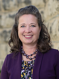 A woman with brown, curled hair wearing a purple beaded necklace, a floral blouse and a purple blazer, smiles for a professional portrait outside a limestone building. 