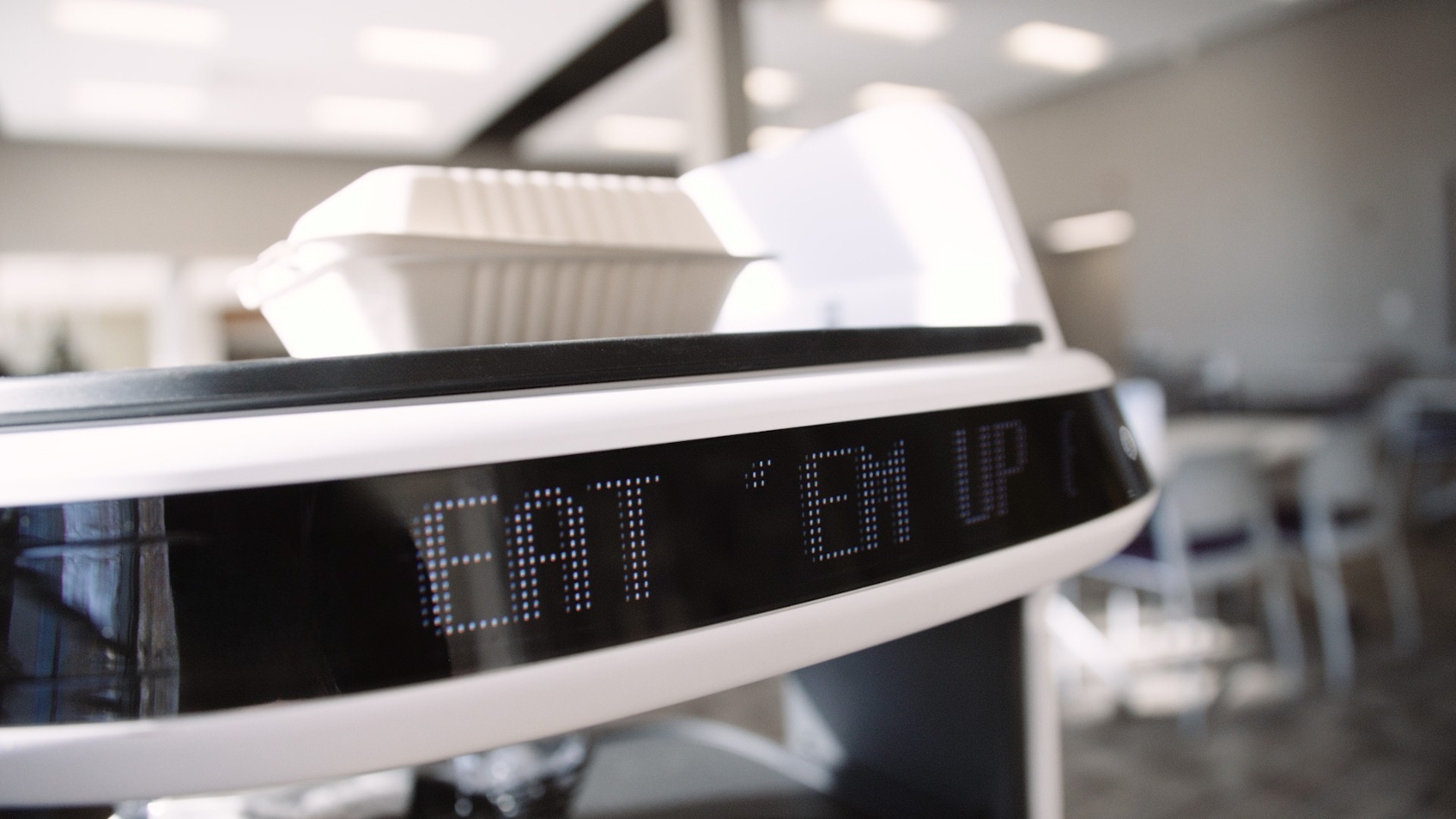 A close-up of the Lacy's food delivery robot displays the message "EAT 'EM UP!" on an LED screen, with a takeout container on top in Hoffman Lounge.