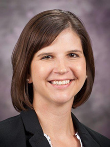 A woman with short, brown hair, wearing a black blazer, smiles for a professional portrait.