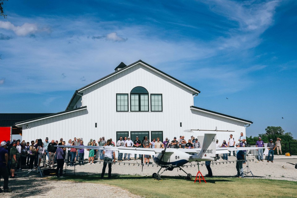 A crowd in front of a wide, white barn watches a man present in front of a large, unmanned aerial plane.