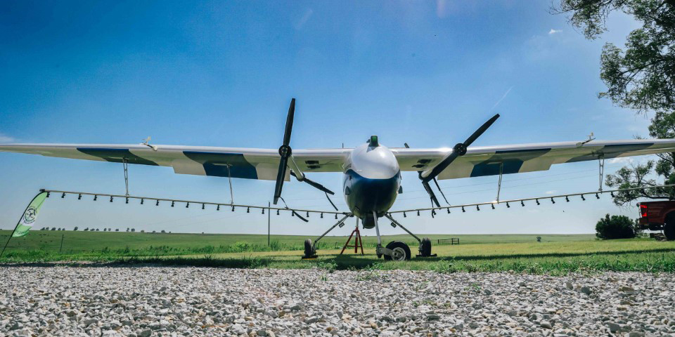 An unmanned, fixed-wing plane flys low across a crop field and sprays it with a chemical.