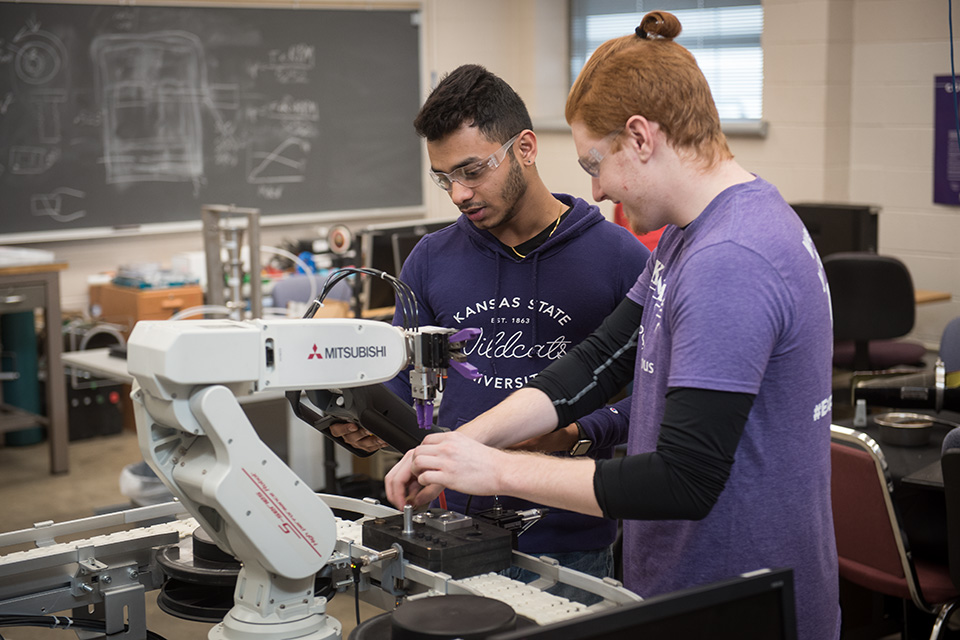 Two Kansas State University students wearing safety glasses work together with a robotic arm and lab equipment during a research activity.