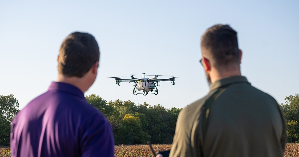 Two men look at and operate an agricultural spraying drone that flies over a field on a sunny, clear day.
