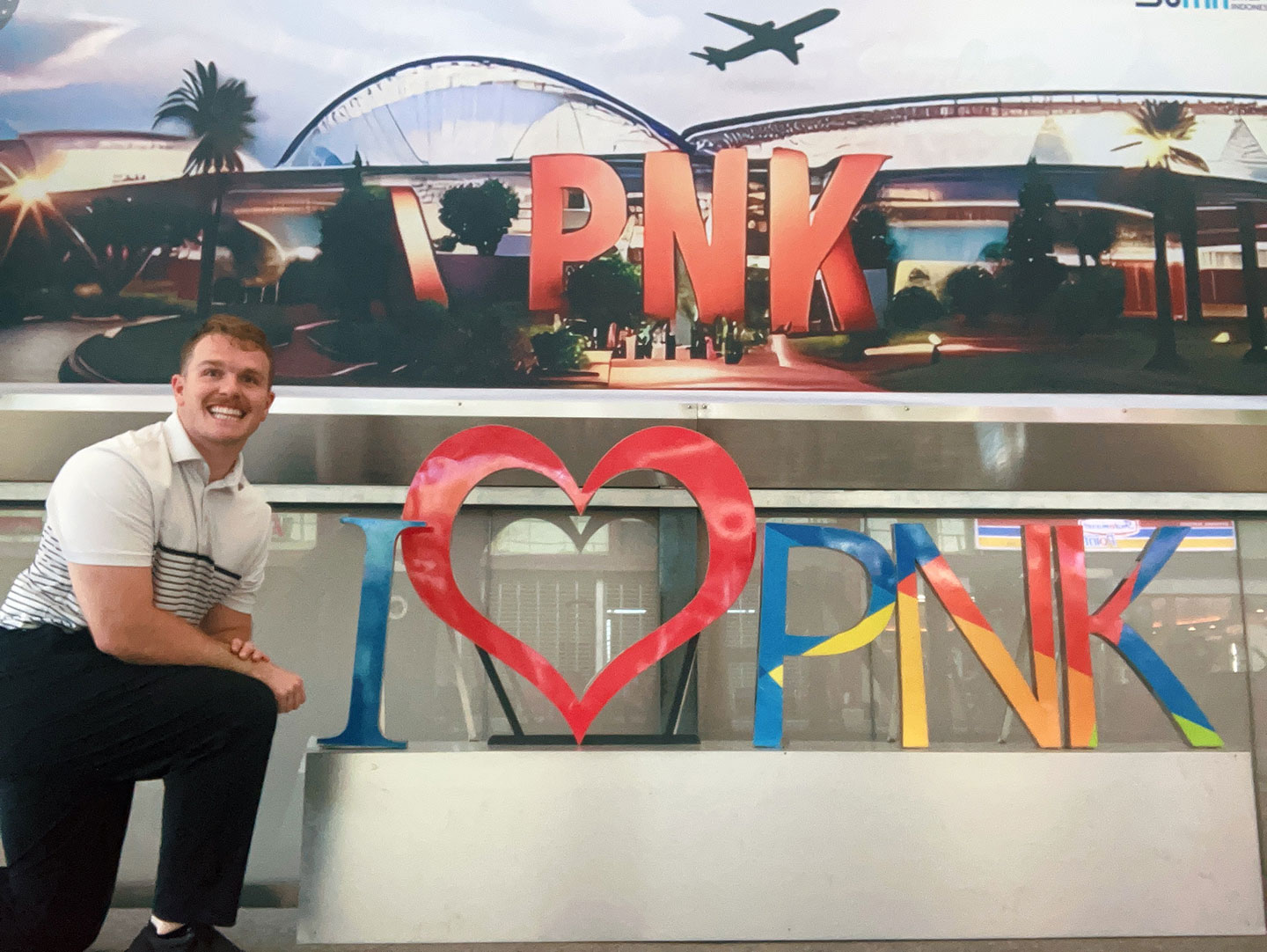 A college student in a white polo stands in front of a sign for the PNK airport.