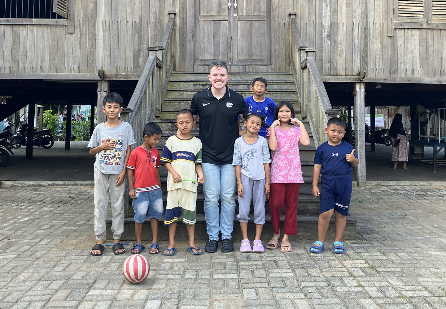 A college student in jeans and a black polo stands with a group of Indonesian children in front of a wooden school.