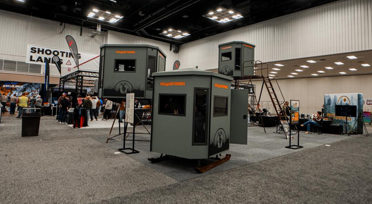 Three hunting blinds with orange text reading RSI OUTDOORS are on display at a trade show. Two of them have stands and are displayed at various heights.