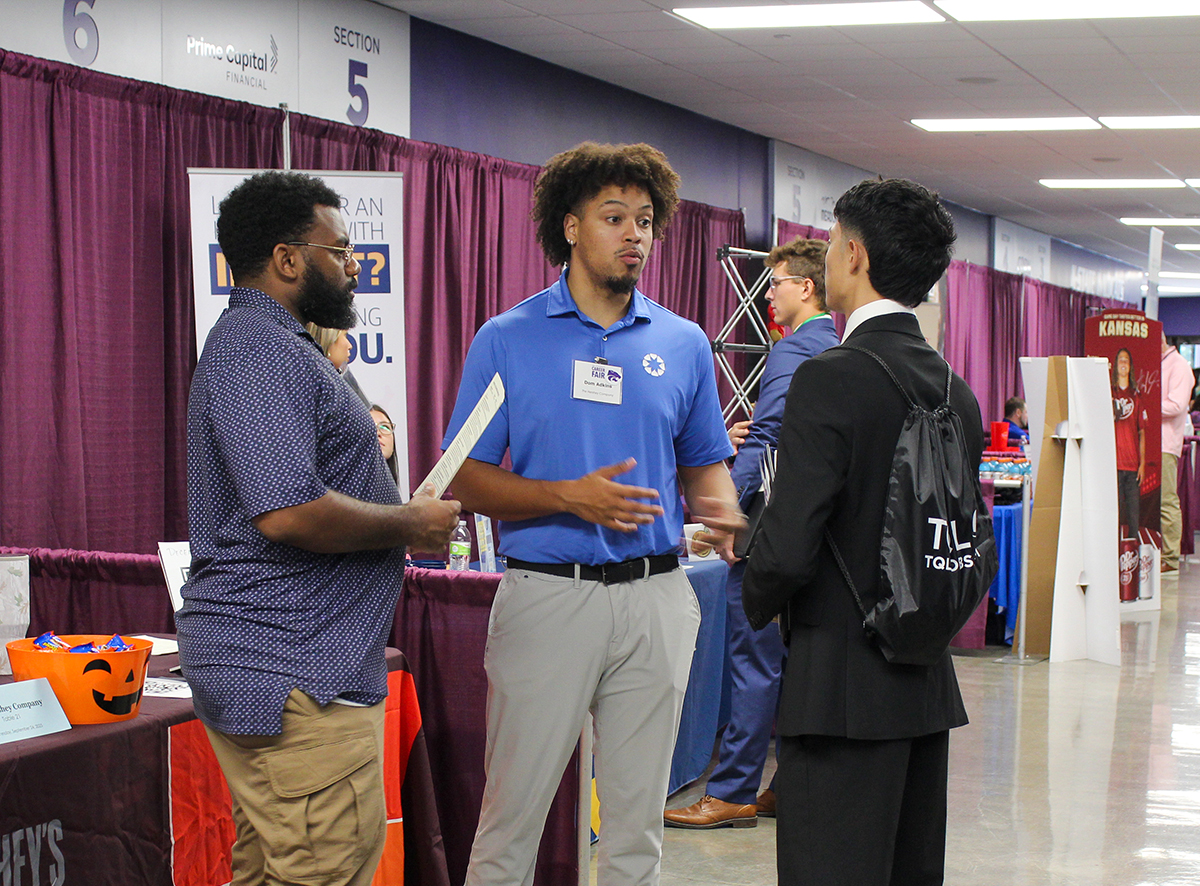 Two Black men in business attire talk to a student in a suit at a university career fair, with tables and decorative backdrops seen throughout the background.