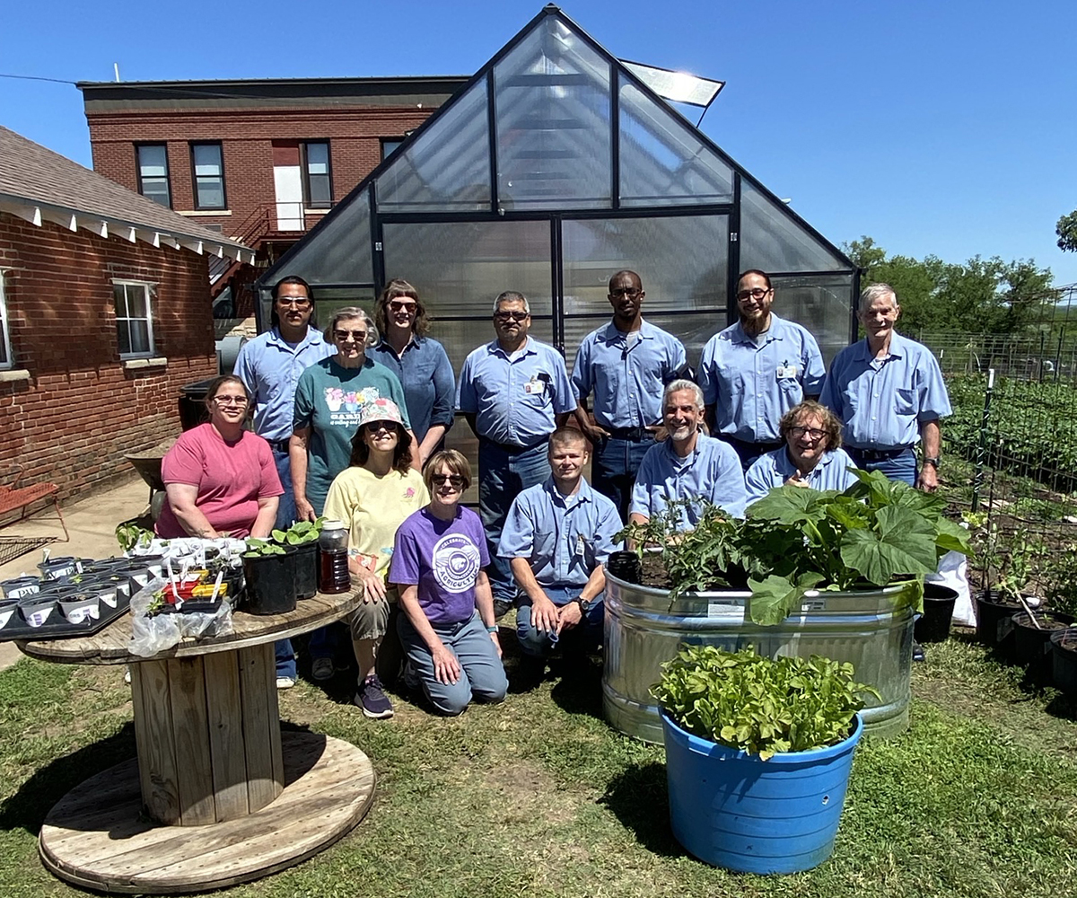 A group of men in light-blue shirts pose alongside volunteers outside of a garden beside a low-security correctional facility.