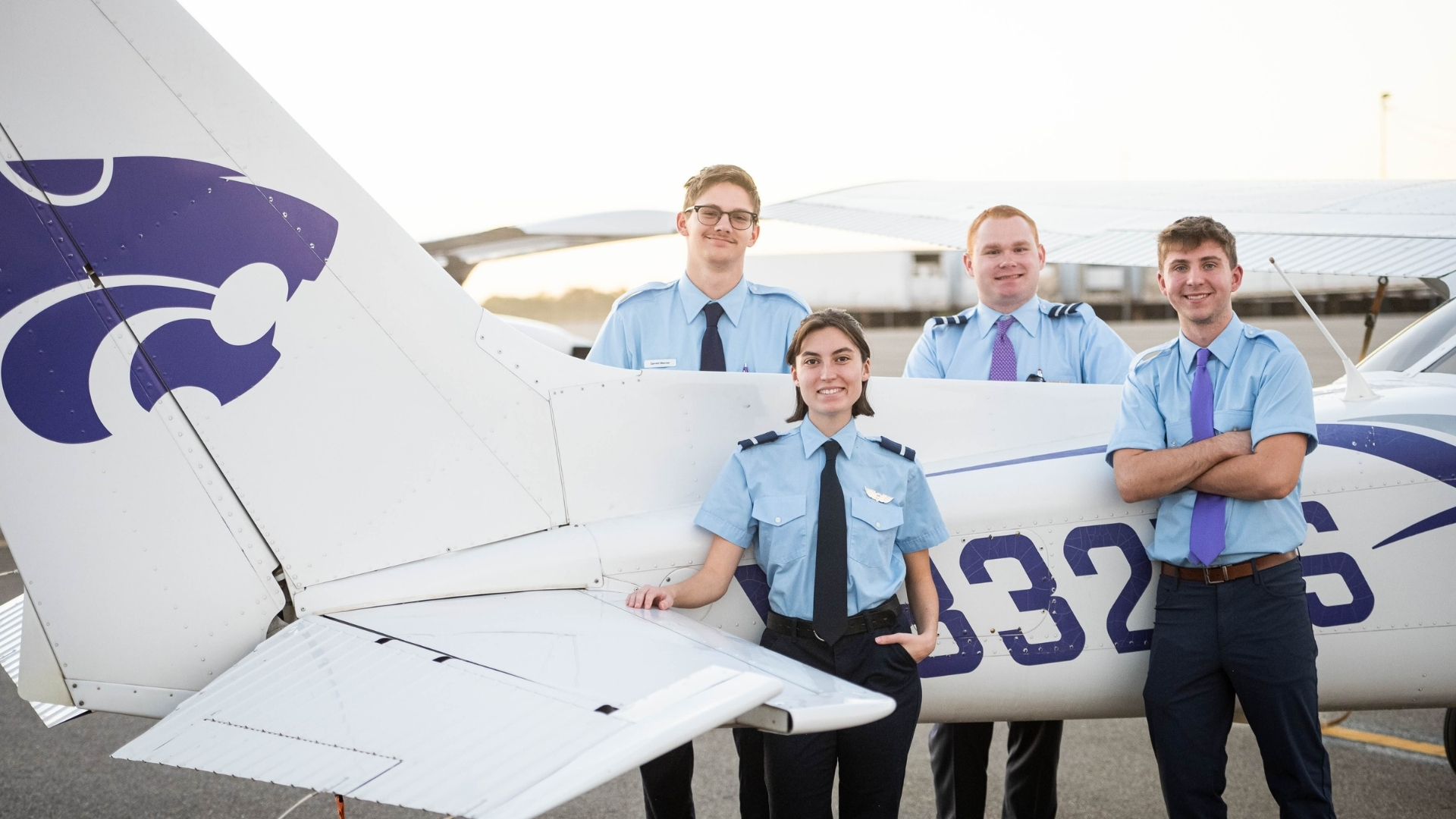 Four young adults wearing light blue pilot-style shirts and ties stand smiling next to a small white airplane with purple markings on the tail at an airport or airfield.