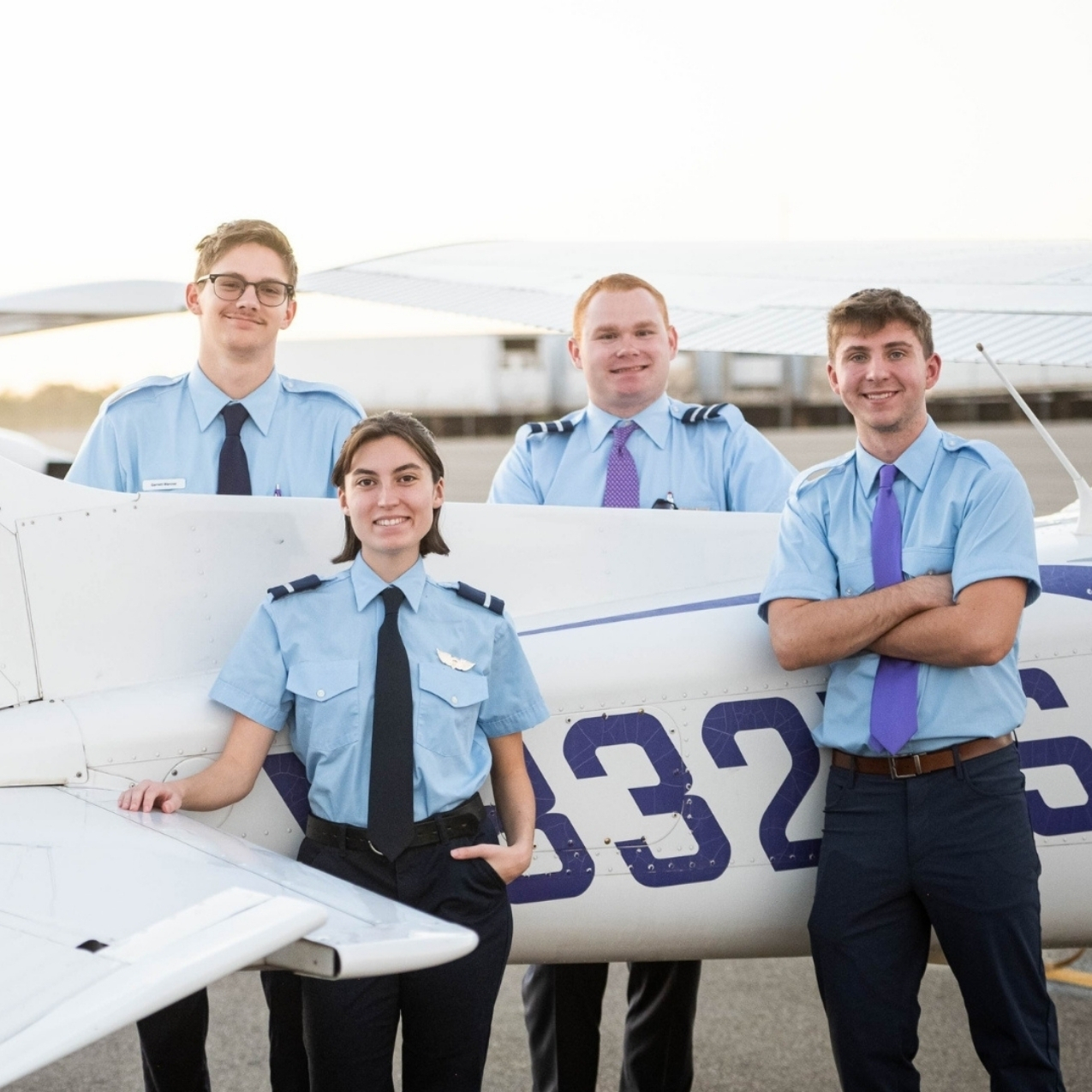 Four young adults wearing light blue pilot-style shirts and ties stand smiling next to a small white airplane with purple markings on the tail at an airport or airfield.