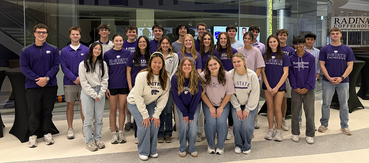 A group of about 30 students, all wearing K-State shirts and either jeans or shorts, stand in three rows in front of a glass wall.