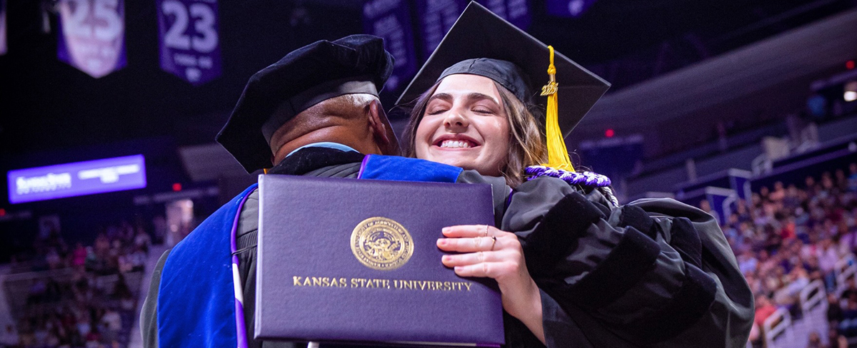 A young woman with brown hair hugs a professor at graduation with her diploma. The professor has a black gown and lea blue stole, and the graduate has a black gown and a purple honor cord on her shoulder.