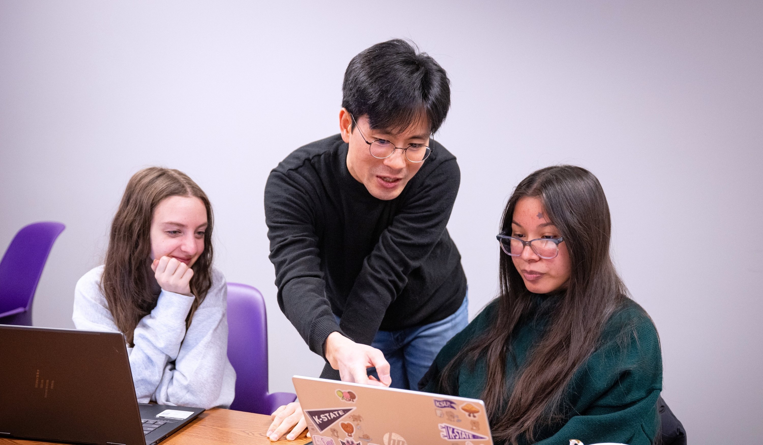 Jin Lee, wearing a black turtleneck and jeans, helps two students sitting at a table with computer work. Standing in the middle and leaning over the table, Lee points to the student's computer on his right. Both students look at the screen attentively.
