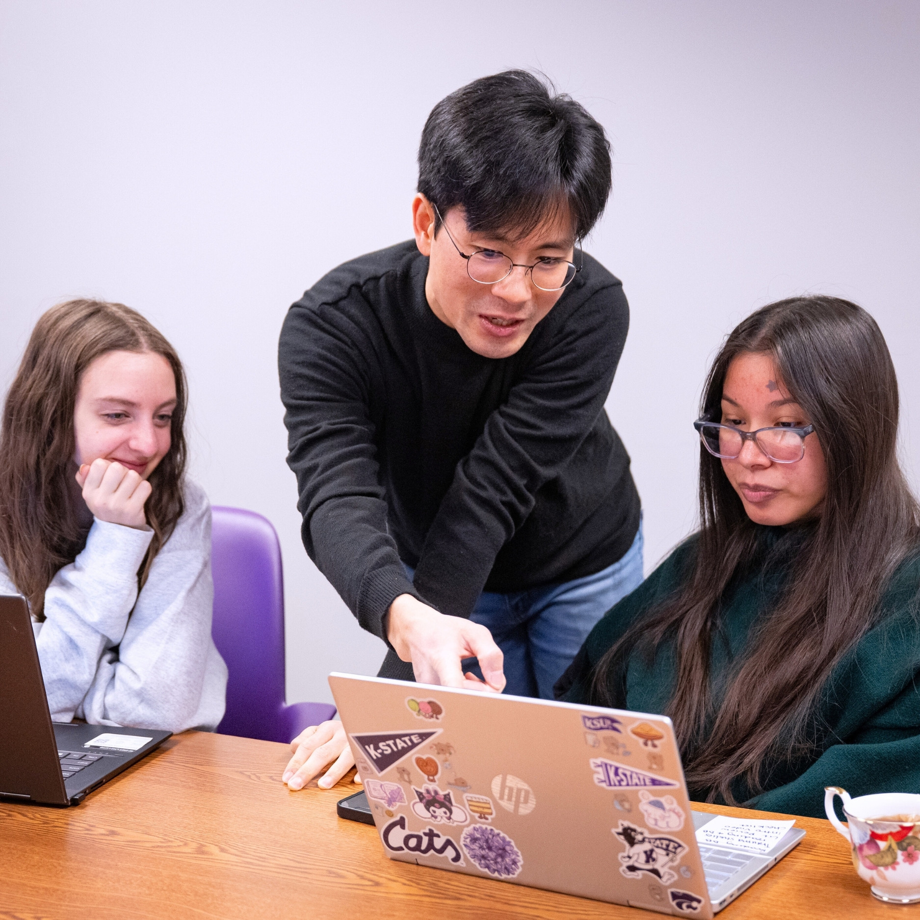 Jin Lee, wearing a black turtleneck and jeans, helps two students sitting at a table with computer work. Standing in the middle and leaning over the table, Lee points to the student's computer on his right. Both students look at the screen attentively.