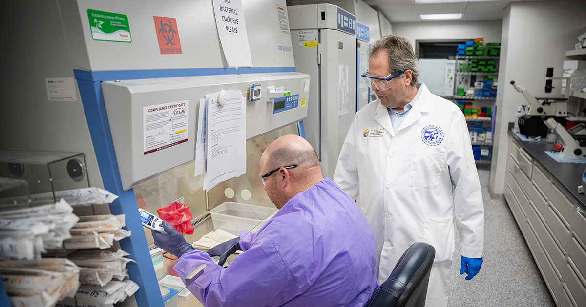 A veterinary researcher in a white lab coat stands over a doctoral researcher in a purple labcoat as the doctoral researcher works with samples under a hood.